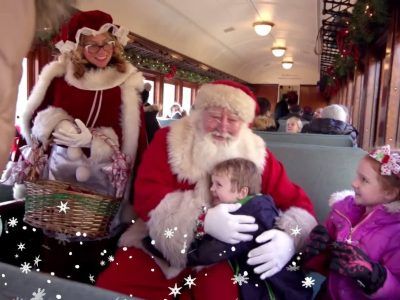 Jultomten kramar ett barn i ett dekorerat tåg, omgiven av frun och ett annat barn.